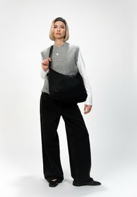 Schwarze Stoff-Schultertasche mit glatter Textur und Reißverschluss. Getragen von einem Model in einem grauen Weste und schwarzen weit geschnittenen Hosen.