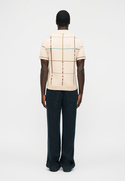 Homme debout, tourné de dos, portant une chemise beige à manches courtes avec des lignes de grille multicolores et un pantalon large et foncé.