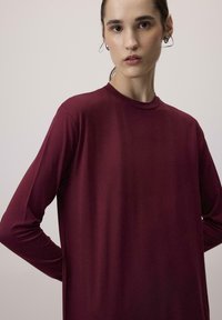Haut à manches longues et col rond, de couleur bordeaux profond, en tissu doux et lisse avec une coupe décontractée. Présente un design minimaliste et un léger éclat subtil.