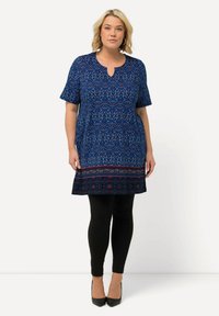 Tunique bleue à manches courtes, motif floral, décolleté en V, ourlet à motifs contrastés, associée à des leggings noirs et des talons noirs.