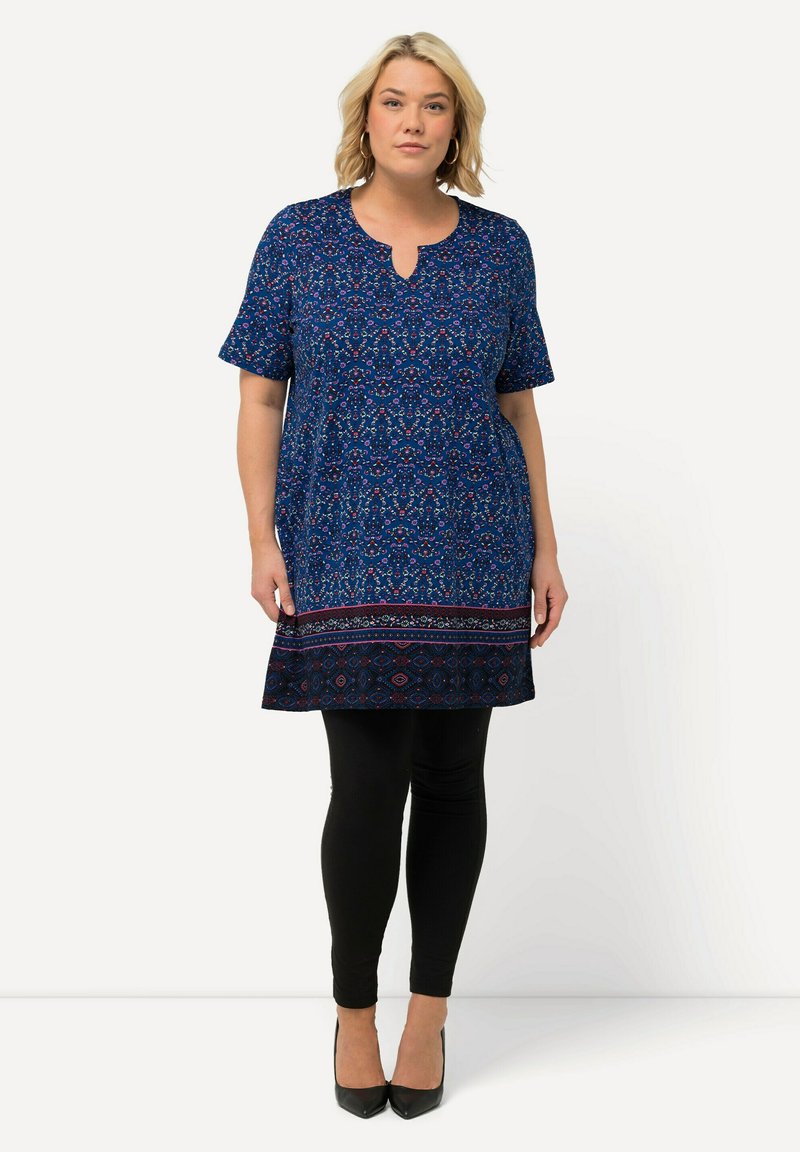 Tunique bleue à manches courtes, motif floral, décolleté en V, ourlet à motifs contrastés, associée à des leggings noirs et des talons noirs.