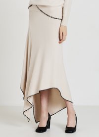 Jupe beige en tricot avec un ourlet haut-bas, ornée d'une bordure noire en forme d'ondulations. Portée avec des chaussures à talons noirs, mettant en valeur la texture et la forme.