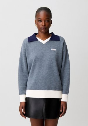 Donna che indossa un maglione grigio con polsini e orlo bianchi, colletto blu navy e una minigonna in pelle nera, in piedi contro uno sfondo semplice.