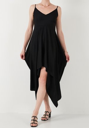 Femme portant une robe noire sans manches à ourlet en mouchoir et des sandales à talons cloutées noires sur un fond blanc uni.