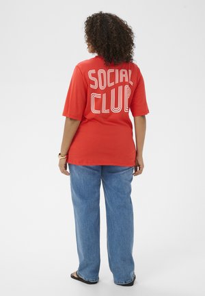Göndör hajú személy piros "SOCIAL CLUB" feliratú pólóban, kék farmerral, szandállal és karórával, háttal áll fehér háttér előtt.