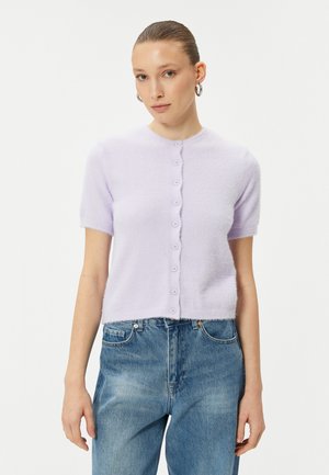 Lavendelkleurige trui met korte mouwen, zachte textuur, ronde halslijn en zeven knopen aan de voorkant; gedragen over een blauwe jeans.