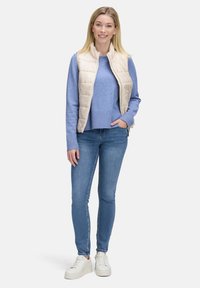 Lekki, kremowy pikowany kamizelka na niebieskim swetrze z prążkowanym wykończeniem, w połączeniu z dopasowanymi niebieskimi dżinsami i białymi butami sportowymi.