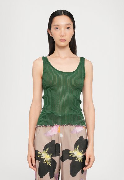 Débardeur vert à côtes avec un col en forme de scoop, présentant un matériau texturé et un ourlet légèrement festonné. Porté avec un pantalon à motif floral.