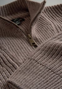 Suéter marrón de punto acanalado con un cuello alto y una cremallera de bronce. Presenta un tejido texturizado y una etiqueta que muestra la talla mediana.