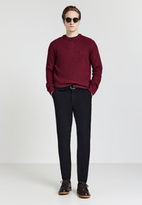 Mann steht mit den Händen in den Taschen, trägt runde Sonnenbrillen, einen burgunderfarbenen Strickpullover, schwarze Hosen und dunkelbraune Schuhe vor einem einfarbigen Hintergrund.