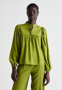 Blusa verde con texture, maniche a sbuffo, un collo alto e chiusura a fiocco. Abbinata a pantaloni verdi coordinati per un look coerente.