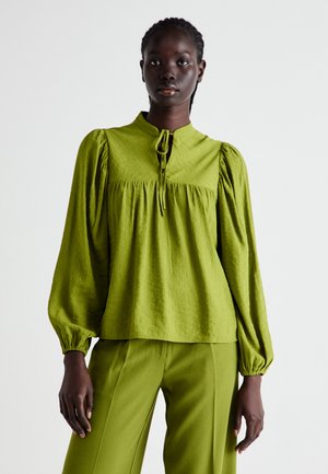 Green textured blouse with puffed sleeves, a high neckline, and a tie closure. Paired with matching green trousers for a cohesive look.
