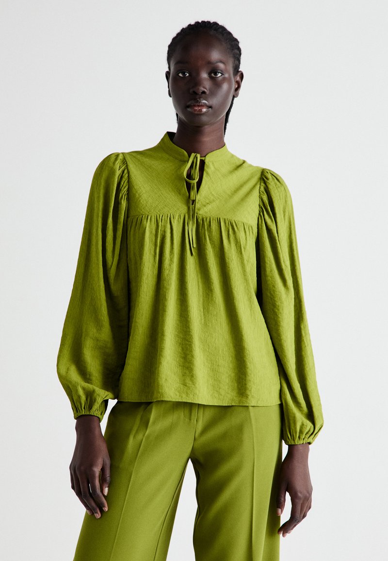 Blusa verde con texture, maniche a sbuffo, un collo alto e chiusura a fiocco. Abbinata a pantaloni verdi coordinati per un look coerente.