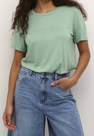 Persoon draagt een lichtgroen t-shirt met geborduurde "Gorgeous" en een high-waisted blauwe spijkerbroek, één hand in de zak.
