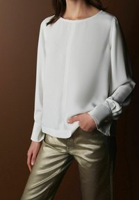 Weißes Langarm-Blusenshirt mit Rundhalsausschnitt, glatter Textur und einfachem Design, kombiniert mit metallischen goldenen Hosen.