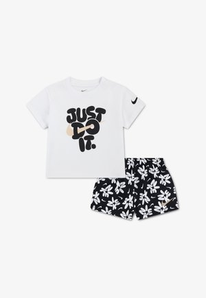 Camiseta blanca de manga corta con el texto "JUST DO IT." y el logo Swoosh, combinada con pantalones cortos negros que presentan patrones florales abstractos blancos y un pequeño Swoosh.