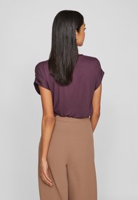 Γυναίκα με ίσια καστανά μαλλιά που φοράει ένα μωβ μπλουζάκι κοντό μανίκι και ανοιχτόχρωμο καφέ παντελόνι ψηλής μέσης, που στέκεται μπροστά σε ένα απλό γκρίζο φόντο.