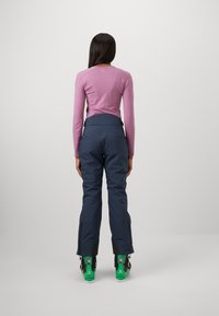 Long-sleeve pink top paired with dark blue snow pants. Green ski boots visible. Pants have a zippered side accent. Smooth fabric texture.