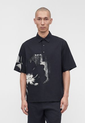 Mand med barberet hoved iført sort kortærmet poloshirt med stort hvidt og gråt blomsterprint, stående foran en ensfarvet hvid baggrund.