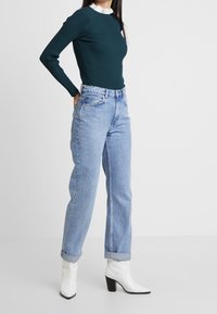 Ciemnozielony prążkowany top z długim rękawem, jasnoniebieskie jeansy z wysokim stanem oraz białe botki na słupku, prezentujące casualową stylizację.