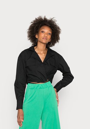 Camisa curta preta com colarinho e dois bolsos no peito, combinada com calças largas de um verde brilhante. O tecido parece suave.