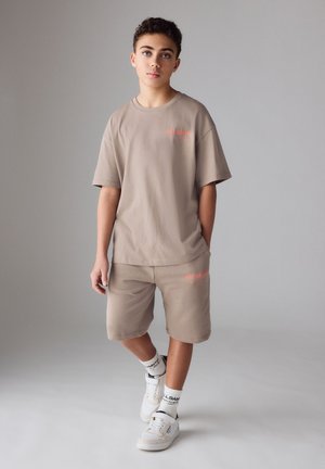 Teismeline poiss seisab käed taskutes, kannab beežikat suurtes mõõtmetes t-särki ning sobivaid lühkse, valgeid sokke ja valgeid tossusid.