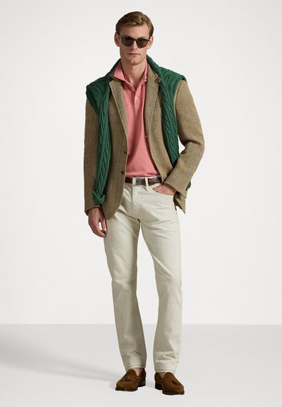 Sacou din tweed maro, cămașă polo roz, pulover tricotat verde purtat pe umeri, pantaloni bej deschis, loafers maro, ochelari de soare negri.