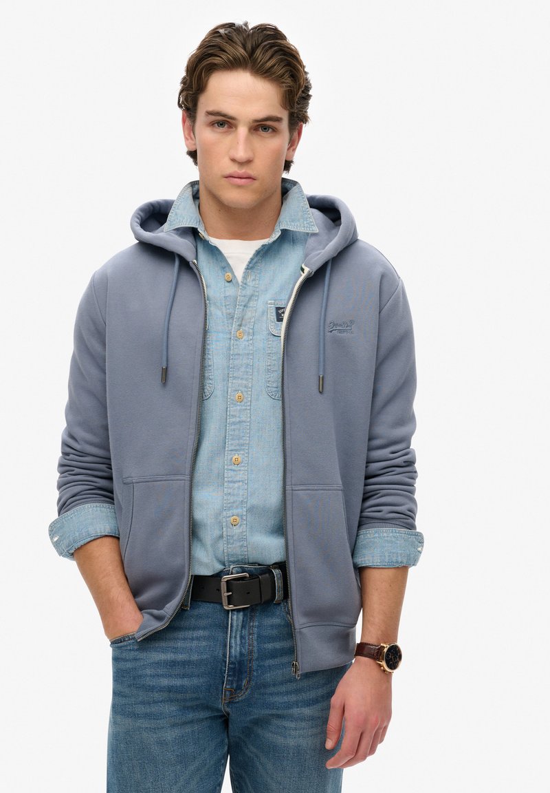 Grijze ritssluiting hoodie met een kangoeroezak over een lichtblauw denimshirt. Donkere jeans en een zwarte riem maken de outfit compleet.