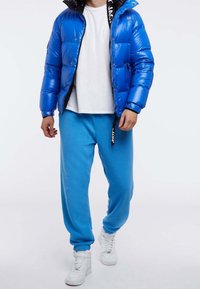 Niebieska kurtka puchowa z połyskującym wykończeniem, zapinana na zamek, z czarnymi akcentami, zestawiona z jasnoniebieskimi spodniami dresowymi i białymi trampkami.