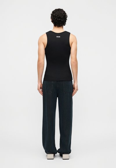 Homme debout, tourné vers l'arrière, portant un haut noir sans manches avec un logo blanc et un pantalon ample foncé, sur un fond clair uni.