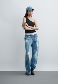 Jeune femme portant un top noir asymétrique, un jean large déchiré, des bottes noires et une casquette en denim bleu, debout, les bras croisés.