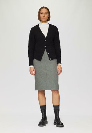 Zwarte gebreide cardigan met knopen, gedragen over een lichte coltrui en een grijze aansluitende rok. Zwarte enkellaarsjes en gestructureerde donkere sokken maken de look compleet.