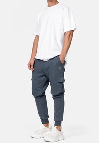 Mężczyzna w białej, oversize'owej koszulce, ciemnoniebieskich spodniach dresowych typu cargo oraz białych, masywnych sneakersach, stojący z rękami w kieszeniach.