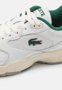 Baskets en cuir blanc avec perforations, doublure intérieure verte, lacets blancs et un logo de crocodile vert sur le côté.