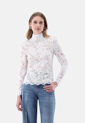 Donna che indossa un top bianco in pizzo a maniche lunghe con motivi floreali abbinato a jeans blu a vita alta, in piedi davanti a uno sfondo semplice.