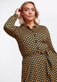 Camicia a maniche lunghe con bottoni nera decorata da un motivo geometrico ripetuto in arancione e verde, con dettaglio in vita annodato.
