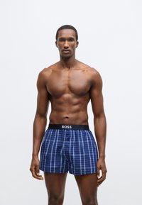 Modri karo boksarji s črnim pasom, na katerem je napis blagovne znamke "BOSS." Tkanina ima gladko teksturo in ohlapen kroj.