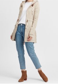 Cárdigan largo de punto color beige con capucha y cordones, combinado con vaqueros azul claro con puños y botines marrones.