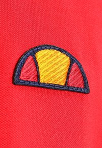 Rødt stof med et blåt broderet mærke, der har et halvcirkelformet design opdelt i gule og røde segmenter.