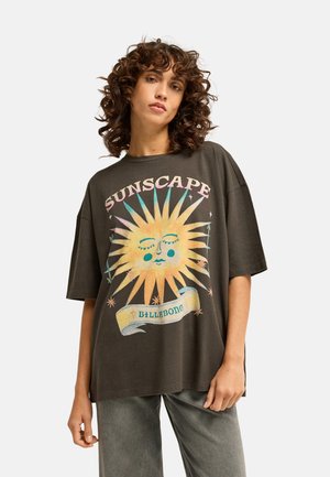 Vrouw met krullend haar die een donker oversized t-shirt draagt met een grote kleurrijke zonafbeelding en de woorden "SUNSCAPE" en "BILLABONG."