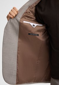 Hellgrauer Blazer mit strukturiertem Äußeren. Braunes Satinfutter mit einer Tasche und einem "Finest Stretch Fabric"-Label. Nahaufnahme des Innenraums.