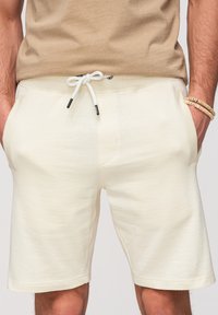 Short beige avec cordon blanc, poches avant, porté par une personne avec les mains dans les poches et une chemise beige rentrée.