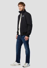 Schwarze Bomberjacke mit Reißverschluss und hohem Kragen, kombiniert mit einem hellgrauen Pullover, blauen Jeans und weißen Sneakers mit grauen Akzenten.