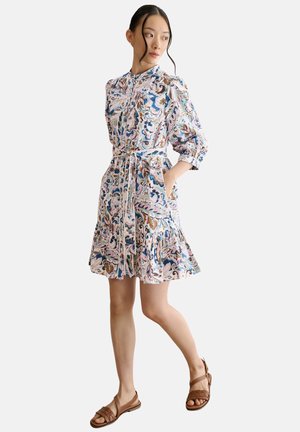 Frau trägt ein pastellfarbenes Kleid mit Blumenmuster, langen Ärmeln und gebundener Taille, kombiniert mit braunen flachen Sandalen, steht vor weißem Hintergrund.