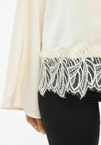 Kremfarget bluse med blondestrim ved nederkanten, med et blomstermønster, kombinert med tettsittende svarte bukser. Glatt stoff, flytende design.