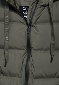 Olivgrüne Steppjacke mit Reißverschluss, ausgestattet mit einem Logo-Label und einem strukturierten Kordelzug. Schwarze Akzente rund um den Reißverschluss sind ebenfalls vorhanden.
