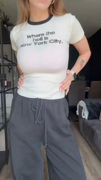Krema majica s črnim obrobo, ki vsebuje drzne črne napise: "Kje za vraga je New York City?" V kombinaciji s svetlo sivo hlačami na vezalke.