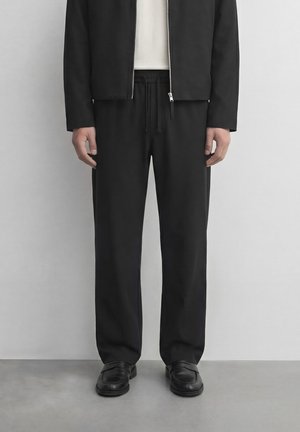 Homme portant une veste noire à fermeture éclair, un pantalon noir avec cordon de serrage, une chemise blanche en dessous et des mocassins en cuir noir, debout sur un sol gris.