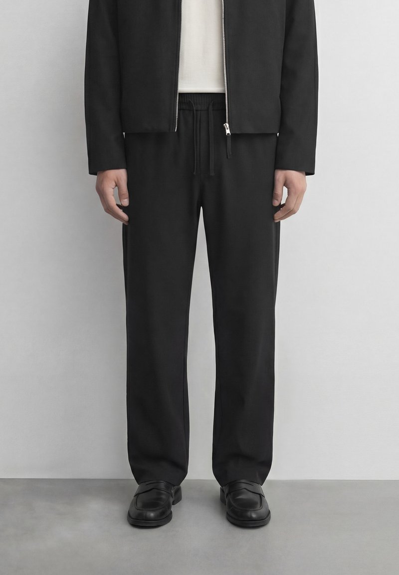 Mand iført sort lynlåstrøje, sorte bukser med snor, hvid skjorte under og sorte læderloafer, står på gråt gulv.