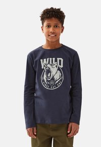 Laivastonsininen pitkähihainen paita, jossa on karhun pään grafiikka ja teksti "WILD SINCE DAY ONE" valkoisena, valmistettu pehmeästä puuvillakankaasta.